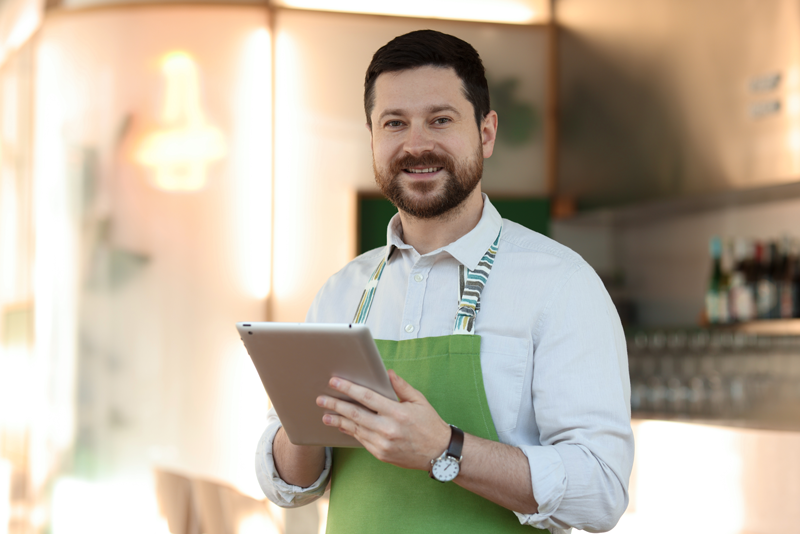 A man wearing a green apron and a light shirt smiles while holding a tablet in a modern, well-lit café, showcasing the welcoming atmosphere of businesses that rely on commercial property insurance in Mississippi.