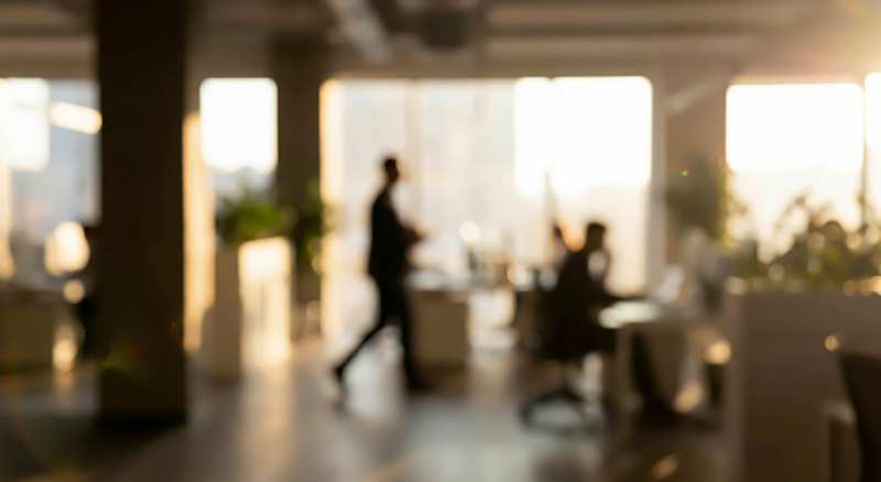 A blurred image of an office with sunlight streaming through large windows, showing indistinct figures of people working at desks and walking—a typical scene in businesses covered by commercial property insurance Mississippi.