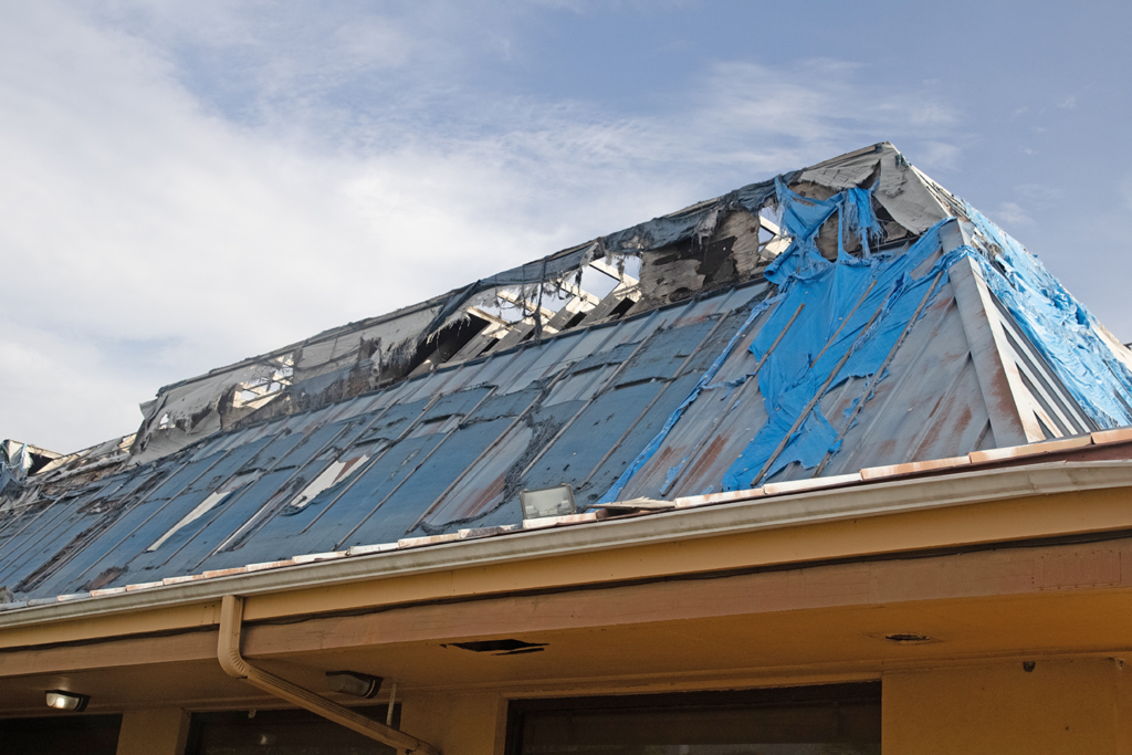 A building’s roof is severely damaged, with missing shingles and a blue tarp covering part of the exposed area—highlighting the importance of commercial property insurance in Mississippi—under a partly cloudy sky.