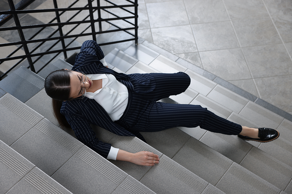 A woman in a pinstripe suit and glasses has fallen while walking up indoor stairs, sitting awkwardly and bracing herself—illustrating why businesses need commercial umbrella liability insurance for unexpected accidents.