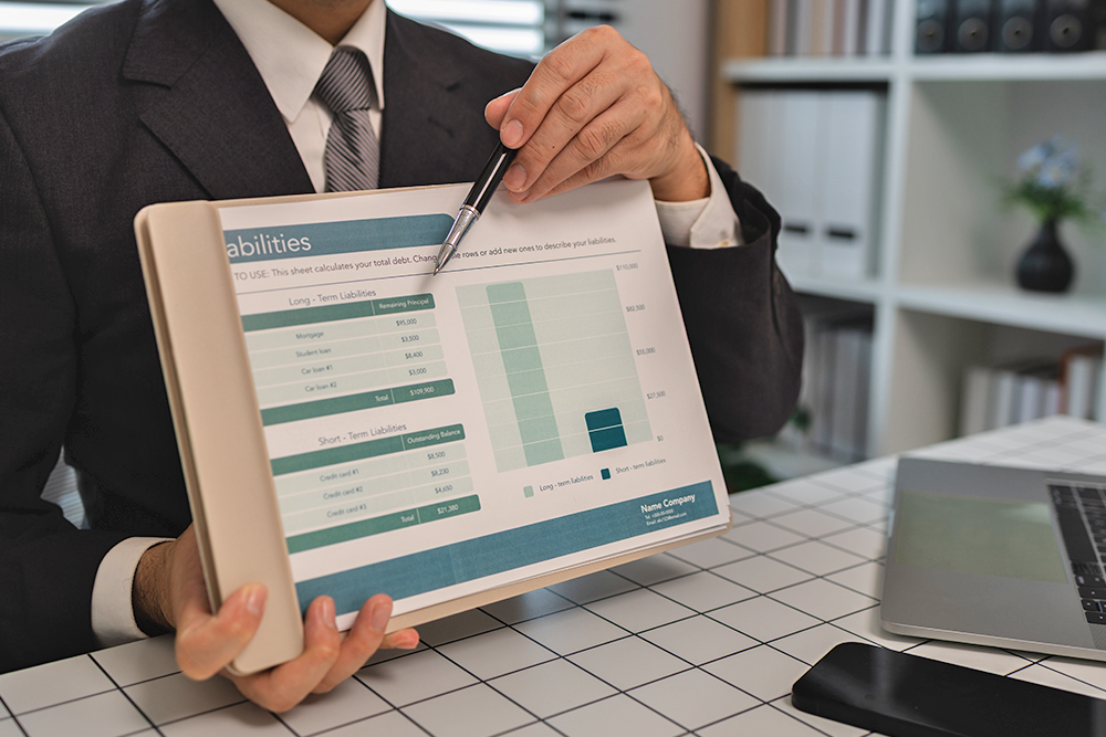 A person in a suit points to a financial liabilities chart on a tablet screen while sitting at a desk with a laptop and documents, illustrating the importance of commercial umbrella liability insurance.