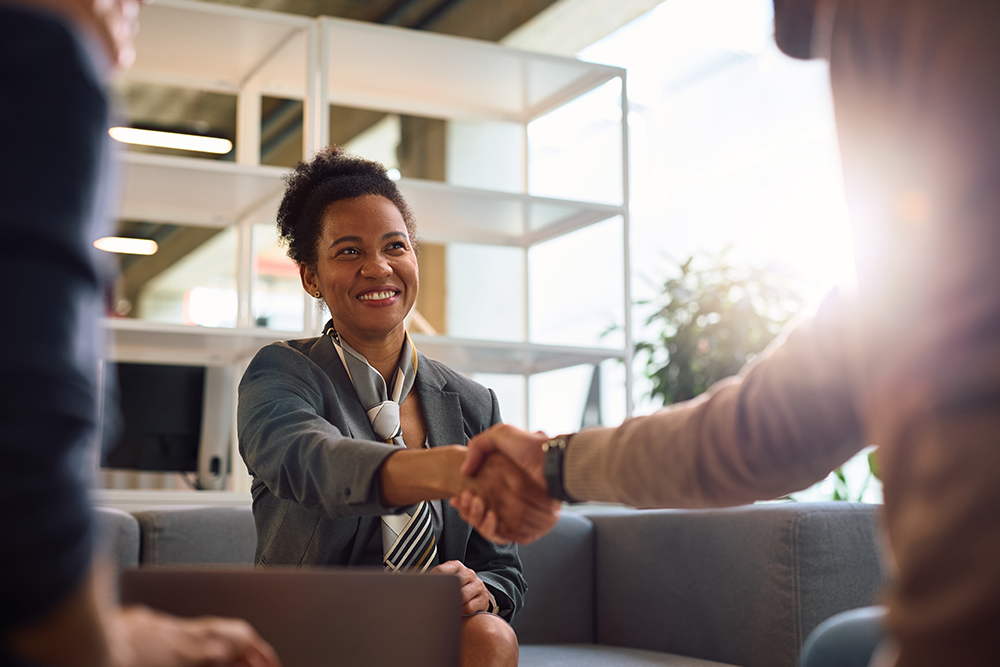 A woman in business attire sits on a couch, smiling and shaking hands with a client, as they discuss commercial umbrella insurance coverage in a modern office setting with shelves and natural light in the background.