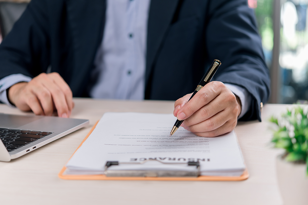 A person in a suit sits at a desk, signing a commercial umbrella insurance document on a clipboard, with a laptop nearby and a small plant visible in the corner.