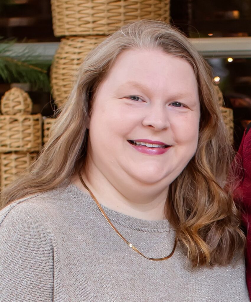 A woman with long, light brown hair is smiling at the camera, wearing a light gray top and a gold necklace. Woven baskets are displayed in the background, creating a warm atmosphere that reflects the inviting style of The James Agency.